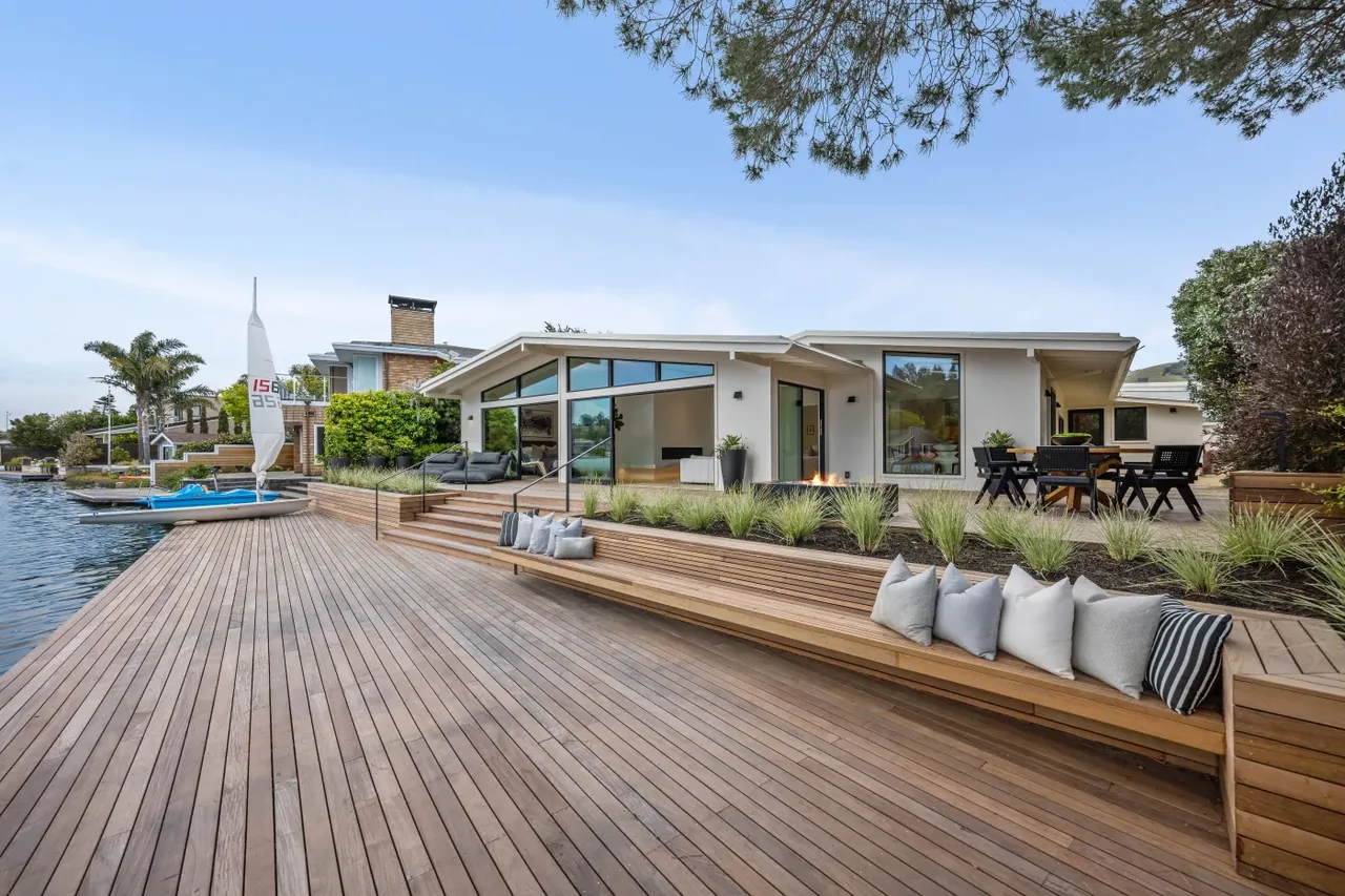 Waterfront deck of a modern home with recessed wooden bench, planter beds, and outdoor dining area shows clean lines and glass walls.