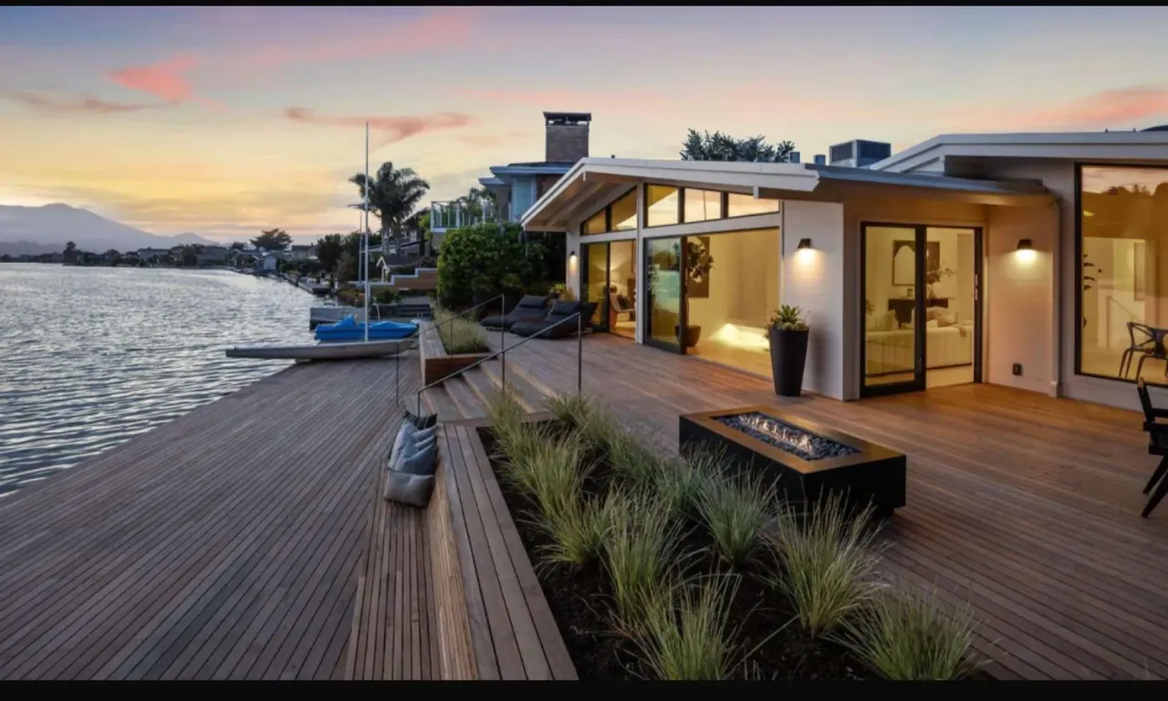 Orla Studios' modern waterfront deck features wood siding, a linear fire pit, and pivot doors opening to the interior.