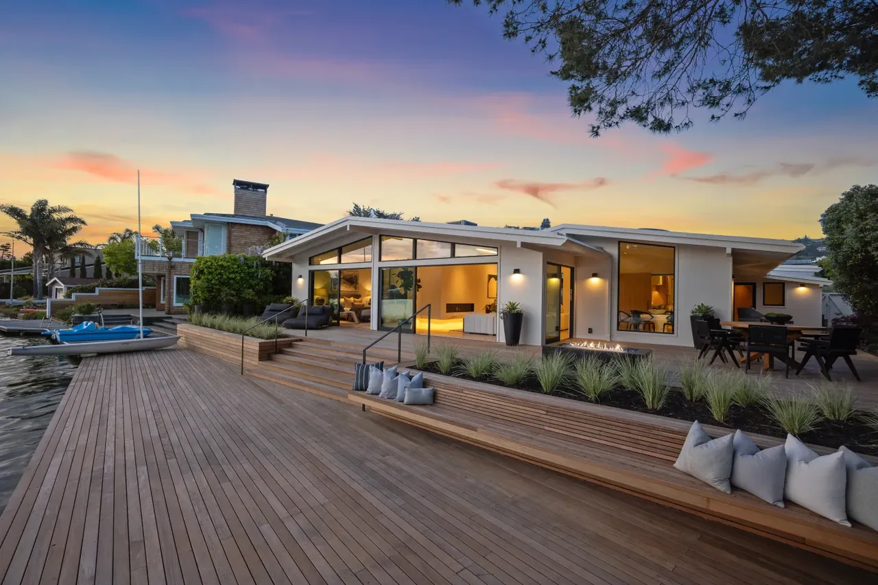 Modern waterfront home with expansive wooden deck featuring planter boxes and outdoor seating at sunset.