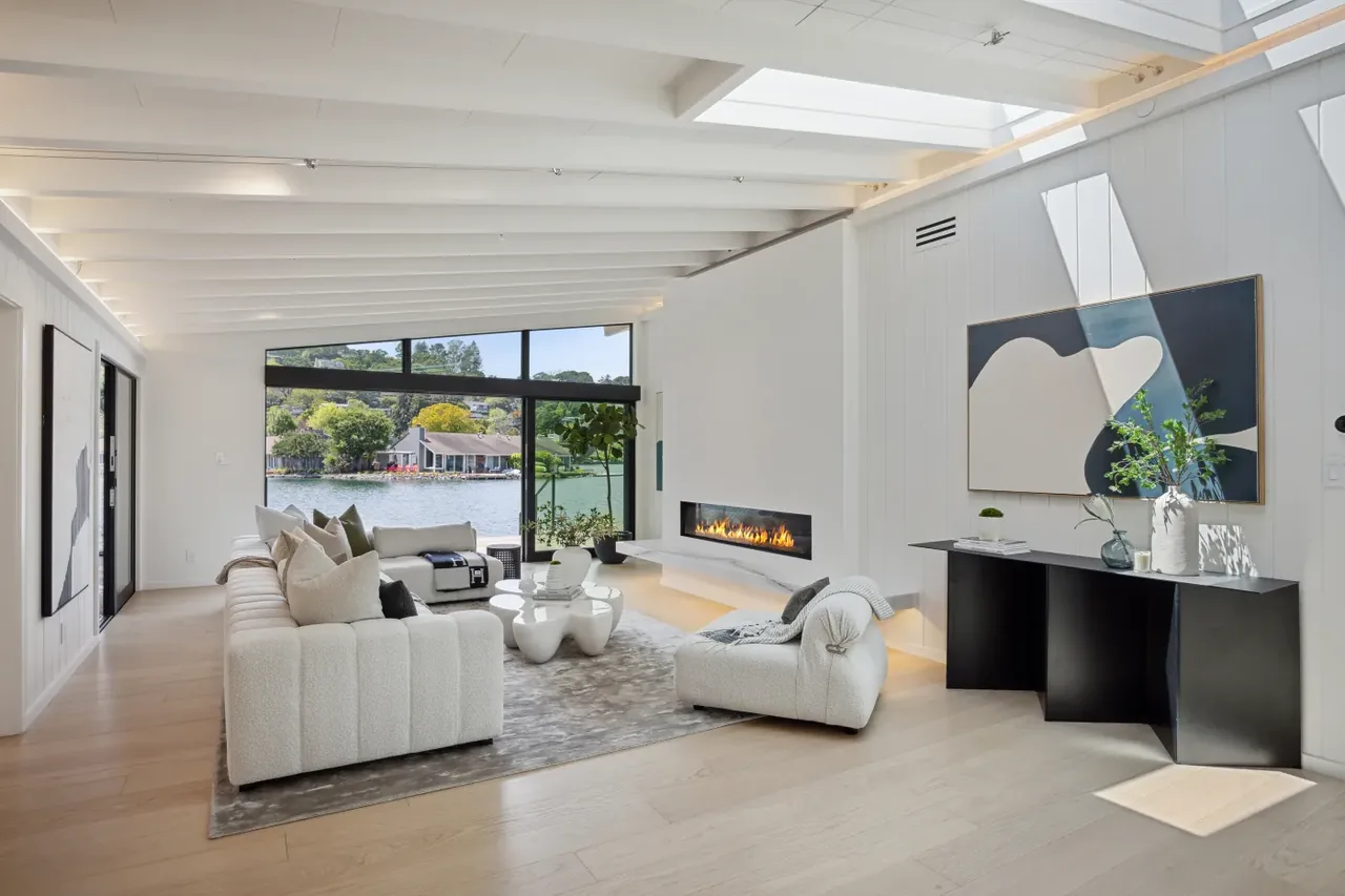 Bright modern living room with exposed ceiling beams, a linear fireplace, and a waterfront view through black-framed windows.