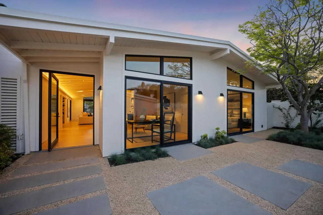 Modern white house exterior with black window frames, pivot doors, and a gravel patio during sunset.
