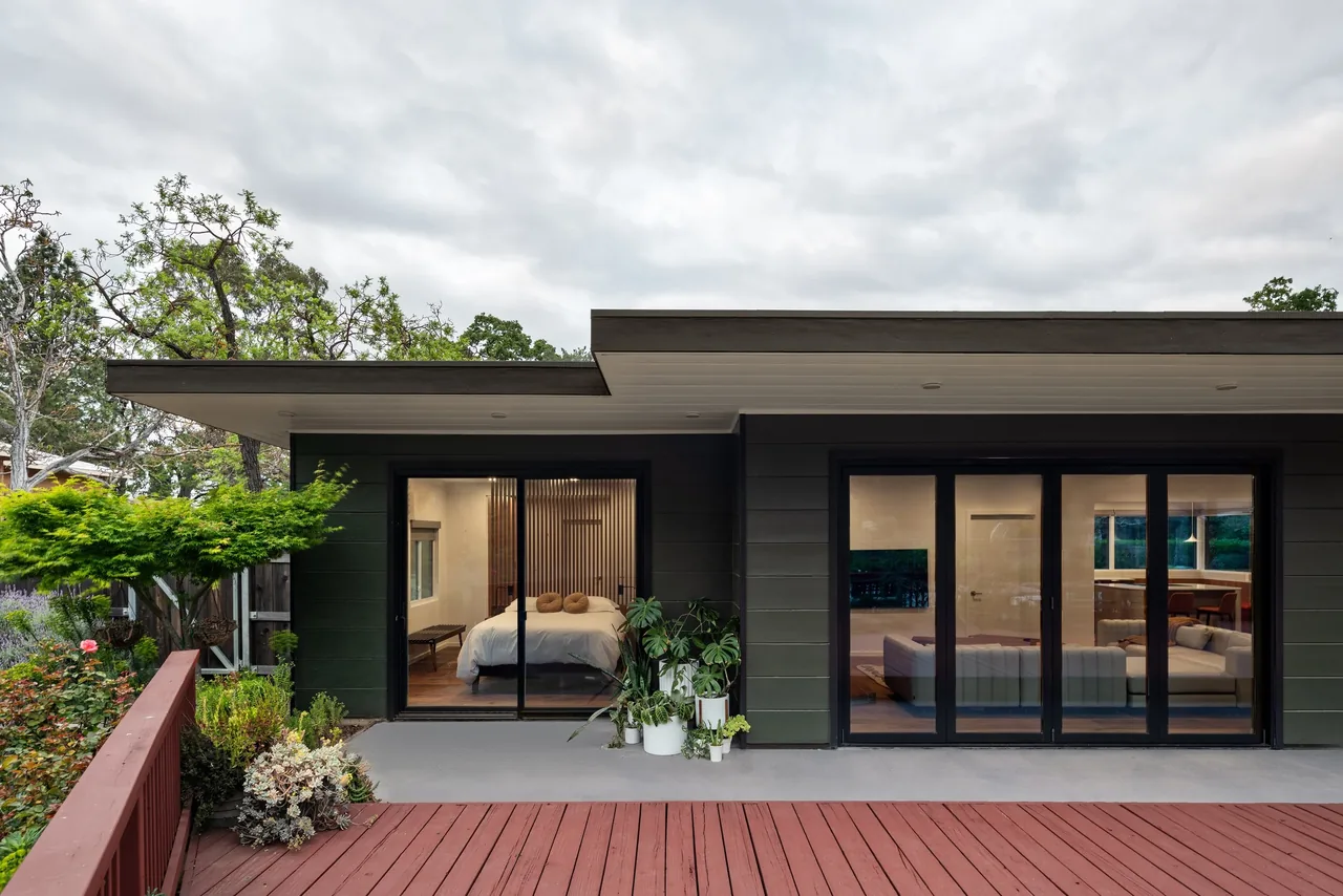 View of a modern flat-roofed house exterior with large black-framed sliding glass doors opening to a bedroom and living room with plants on