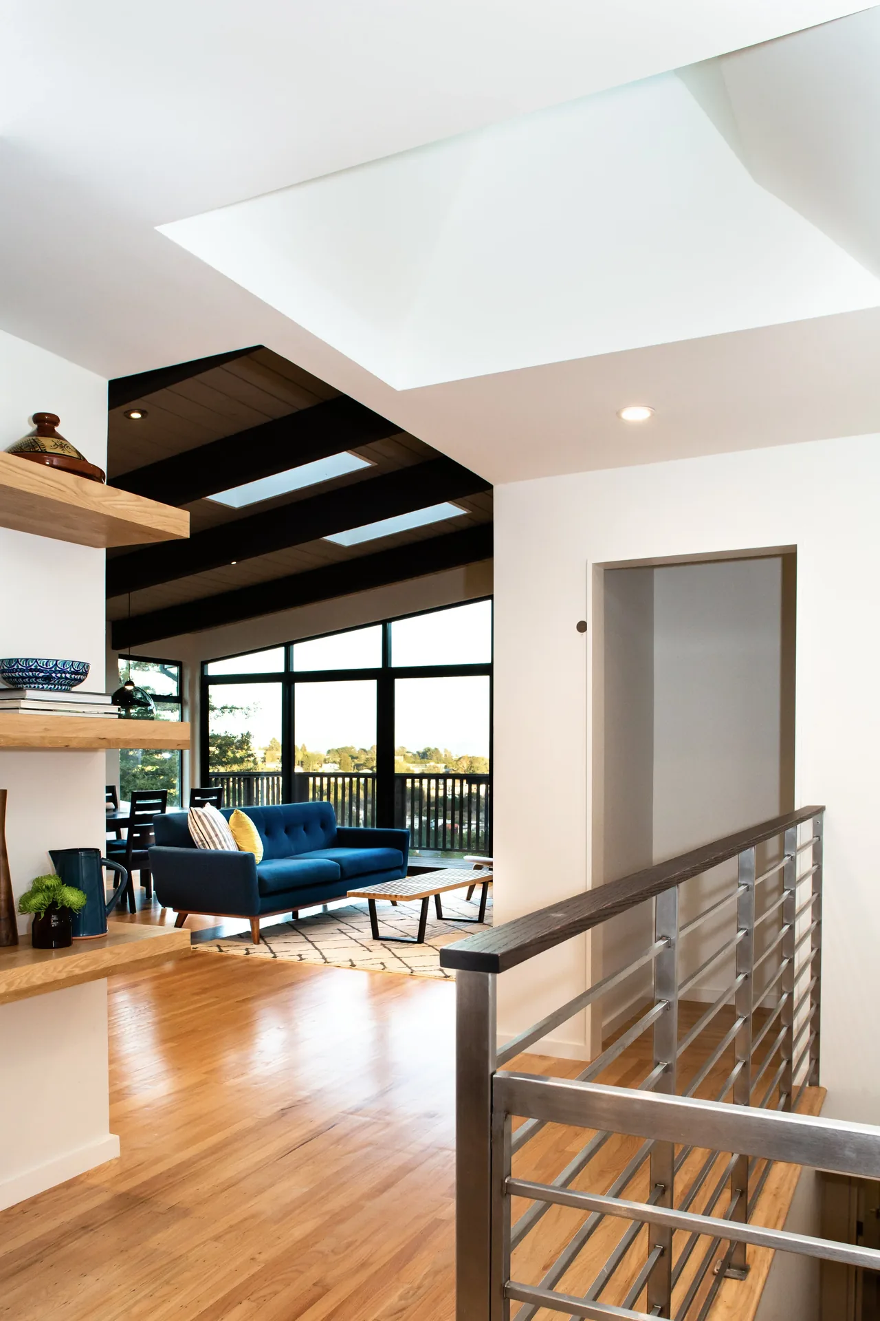 Living area features floor-to-ceiling black-framed windows with views, dark ceiling beams with skylights, and a metal-rimmed railing.