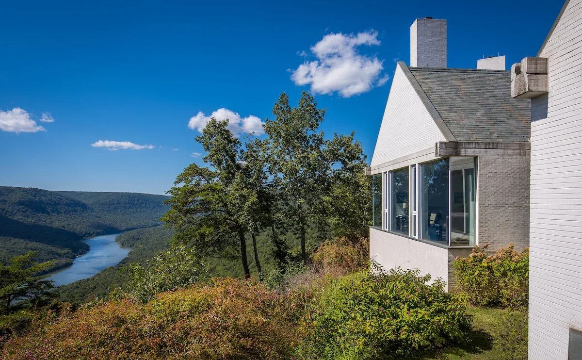 The Mills Residence features a white brick exterior with a tall rectangular chimney and large windows facing a river and wooded hills.