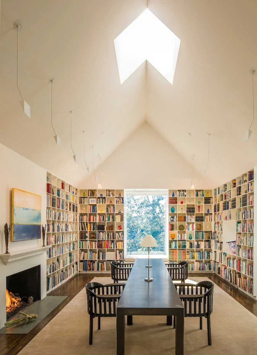 Interior library space featuring vaulted ceiling with skylight, built-in tall bookshelves, a fireplace, and a centered table with chairs.