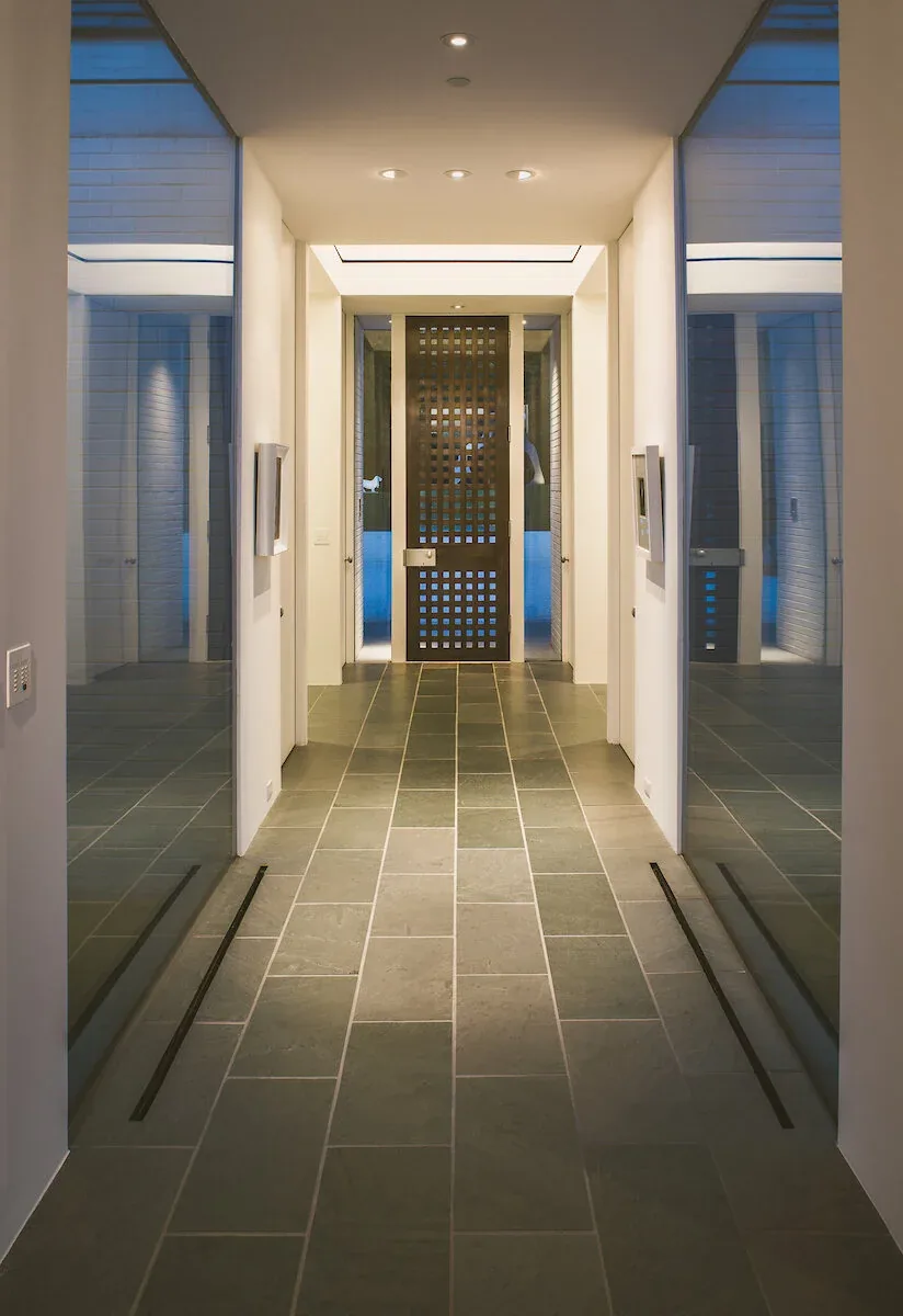 The Mills Residence features a corridor leading to a metal lattice front door with recessed lighting overhead.
