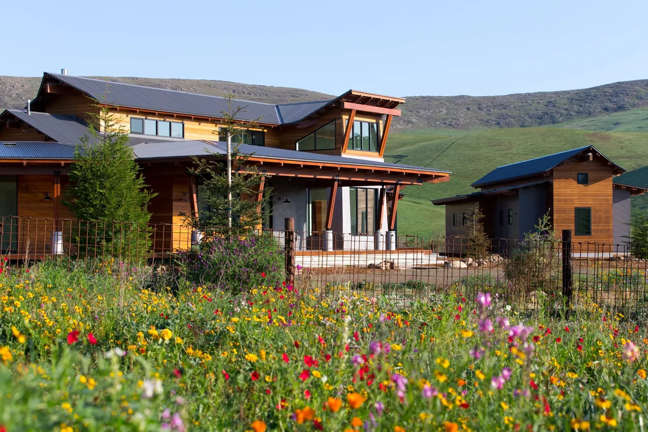 Cayucos Ranch features a wood-sided home with black metal roofs and angled wood braces, nestled in a wildflower meadow and hillside landscape.