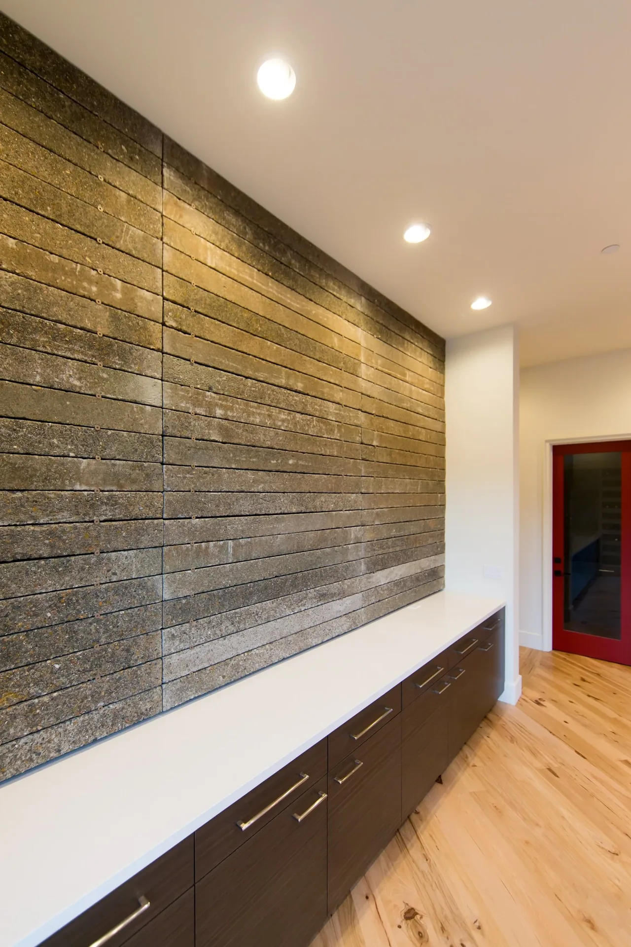 A textured horizontal concrete wall above a white countertop with dark wood cabinets and recessed ceiling lights.