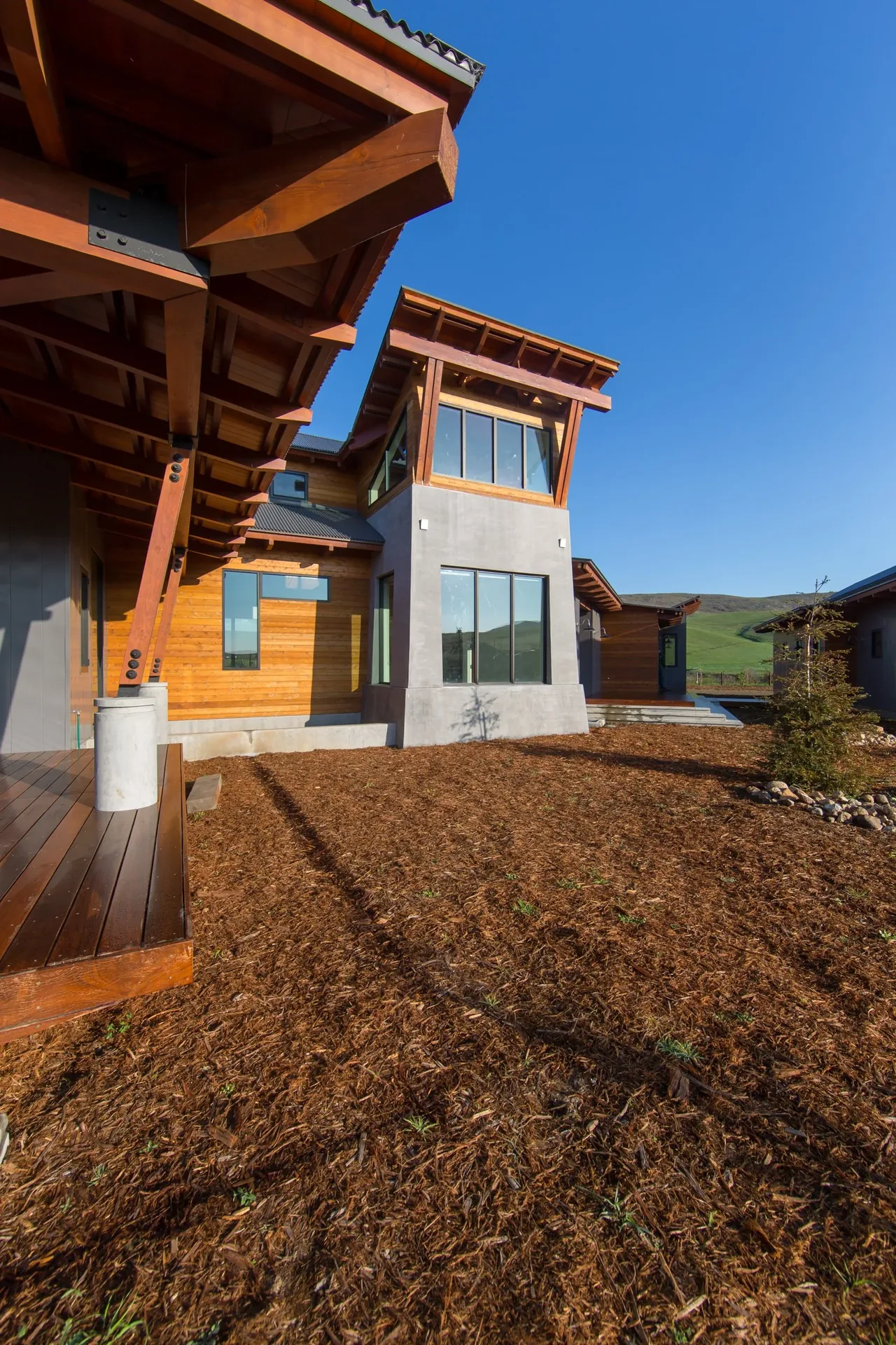 Modern ranch house at Cayucos Ranch featuring wood siding, concrete base, and large-framed windows with a backdrop of rolling green hills.
