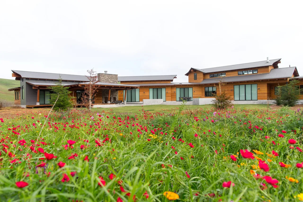 Modern ranch home with wood and metal siding, multiple rooflines, and large glass doors opening to a landscaped yard with wildflowers.