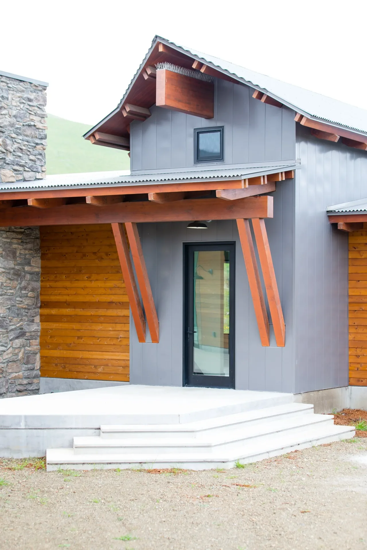 Entry exterior featuring stone, wood siding, gray metal panels, and a gable roof with exposed wood beams at Cayucos Ranch.