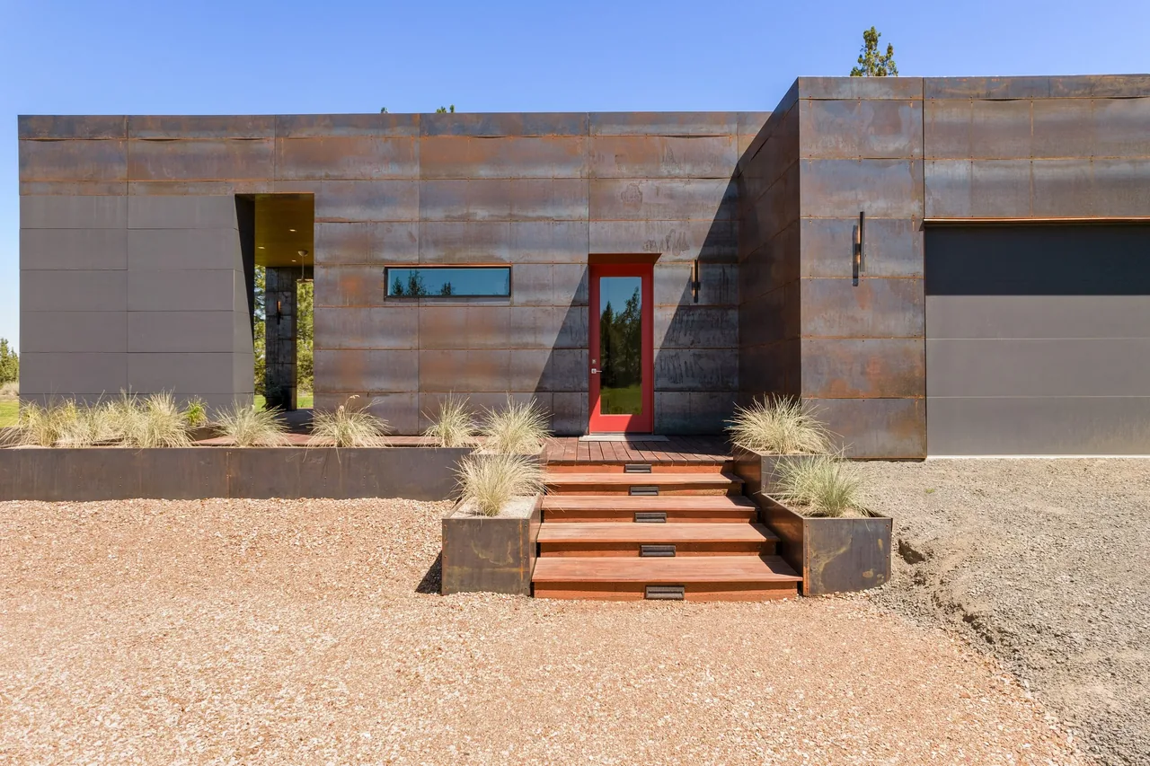 Schibel Road Residence features rusted metal panel facade, red entry door, wooden stair steps, and gravel driveway.