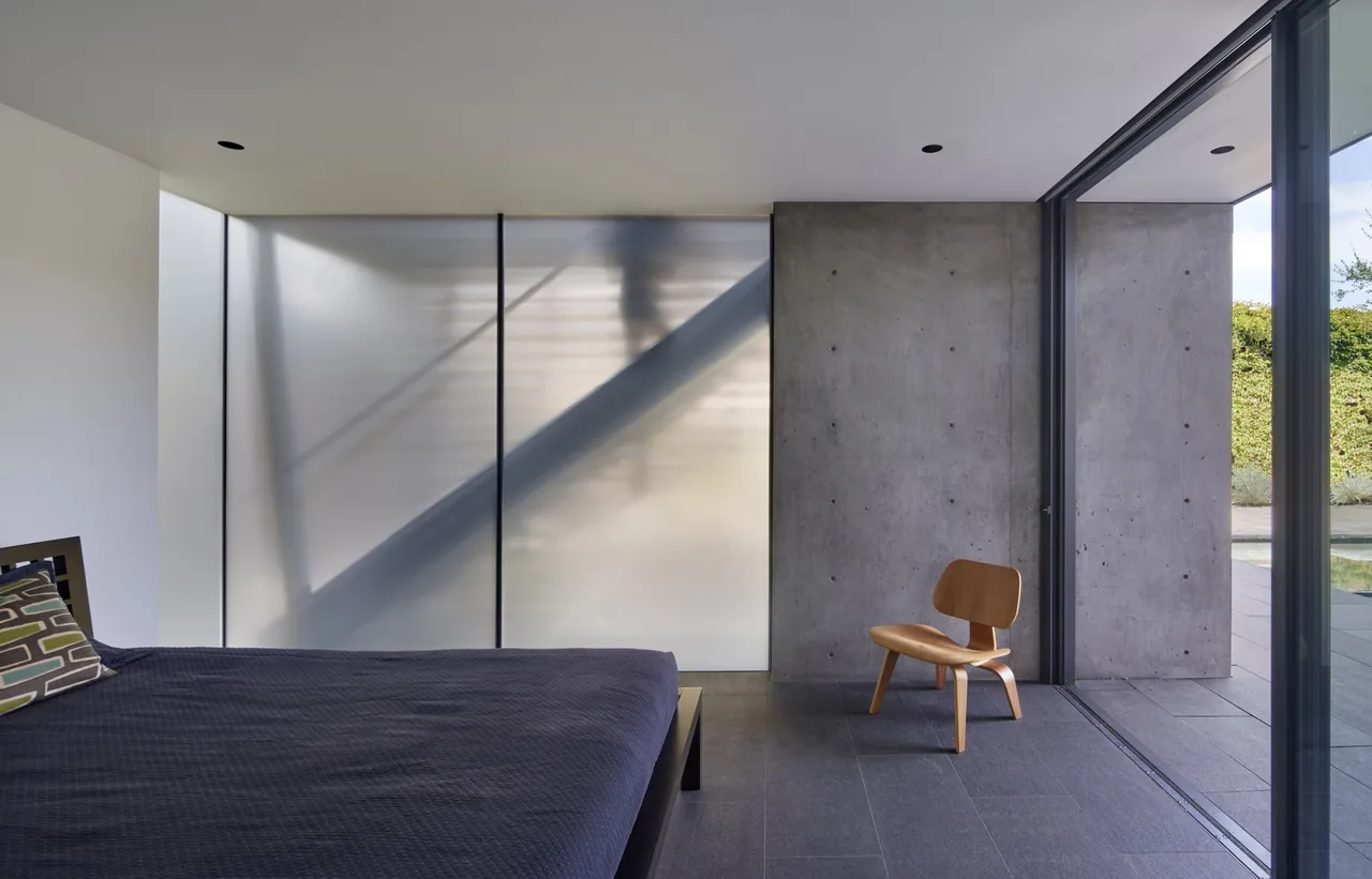 The Edgewood House bedroom features a frosted glass wall, concrete finishes, and expansive floor-to-ceiling windows for seamless indoor-outdoor flow.