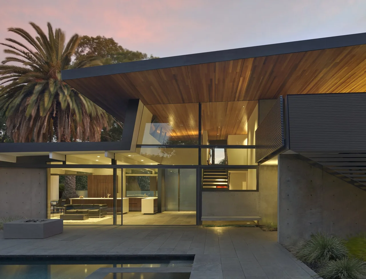 Edgewood House features a cantilevered wood-paneled roof, large glass walls, and a poolside patio with gray stone tile flooring.