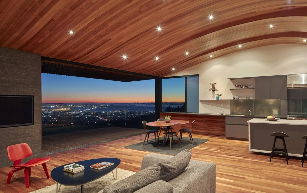 Open-plan living and kitchen area features a curved wood ceiling and expansive opening with glass railing overlooking city views at sunset.