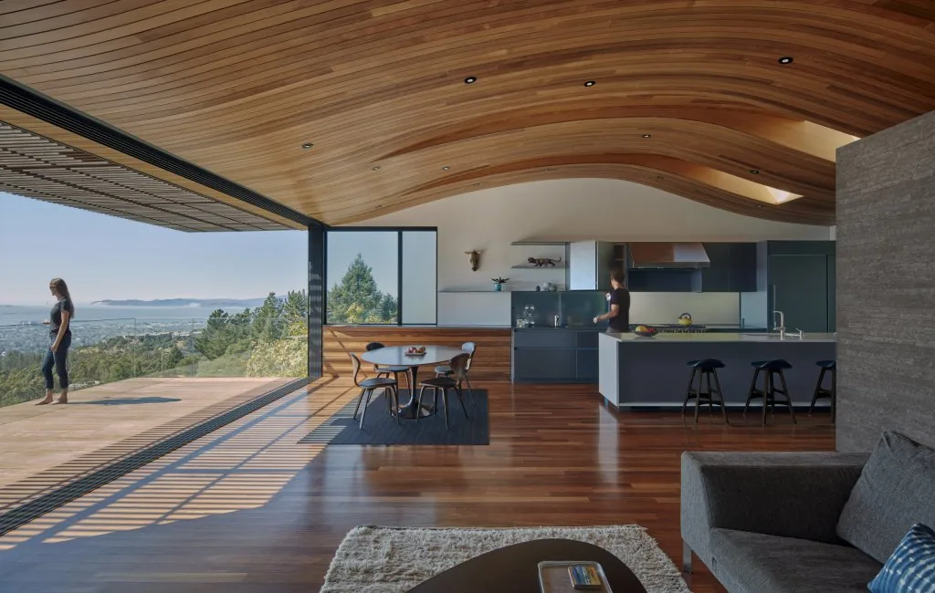 Skyline House features an open-plan living and kitchen with a distinctive curved wood ceiling and a large opening to the deck overlooking the landscape.
