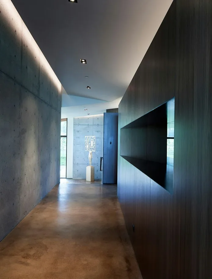 Interior hallway in Diagonal House with polished concrete floor and concrete wall, featuring recessed ceiling lights and dark wood panels.