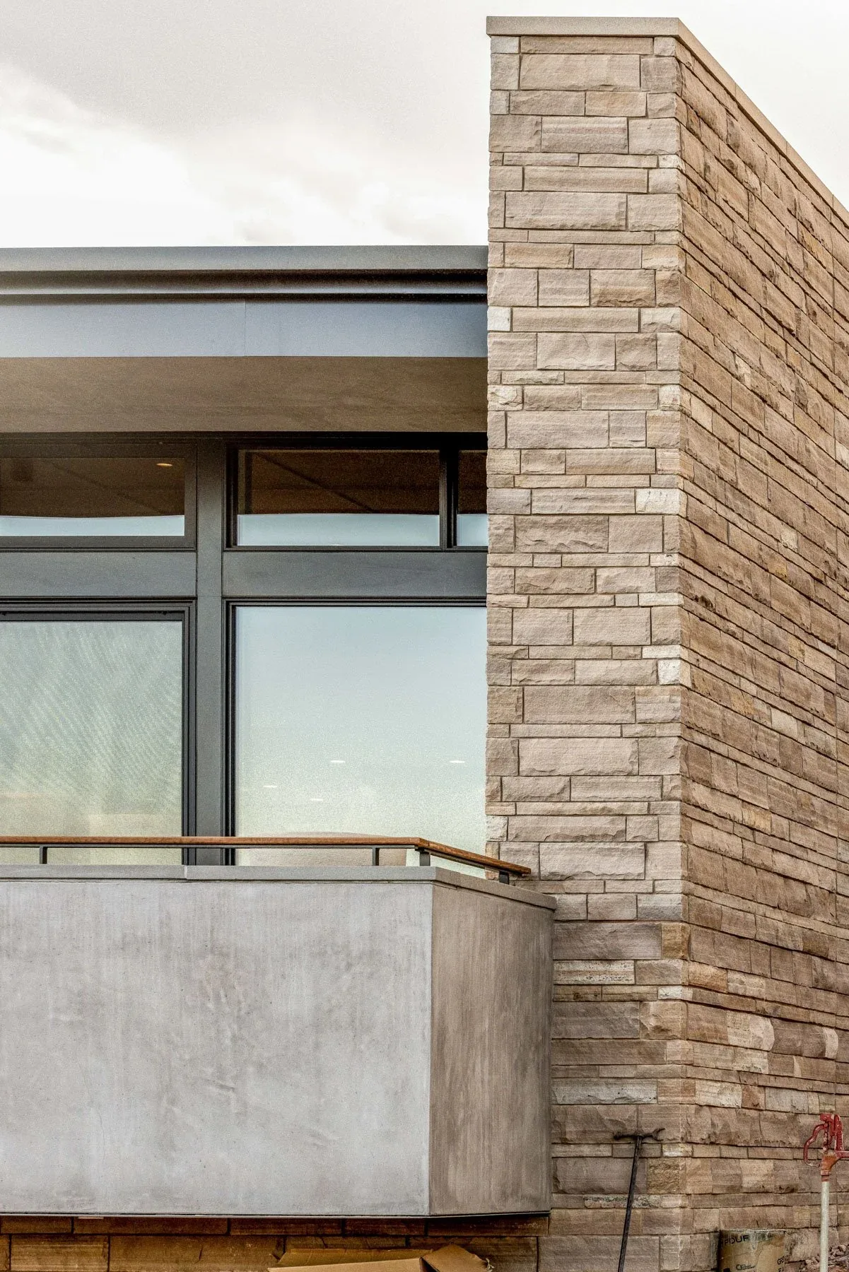 Modern house exterior featuring stone masonry, black window frames, and a concrete balcony with wooden handrail.