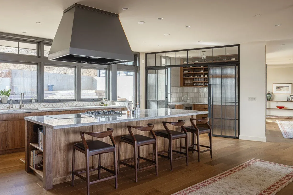 Kitchen with a large wooden island includes a cooktop and seating with dark wood stools and large windows above the main counter.