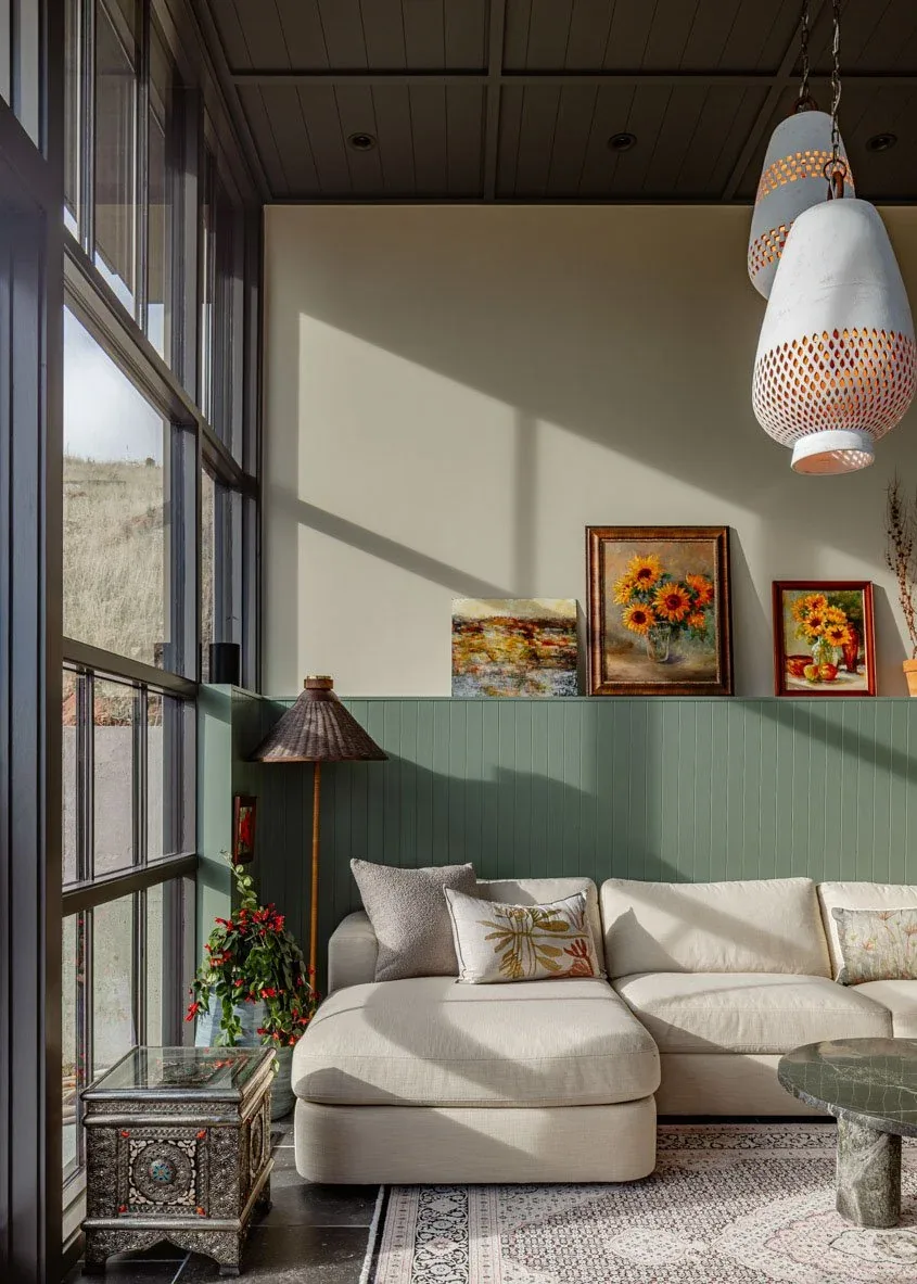 Living room in Boulder Mountain Residence shows gray window frames, beige sofa, green wainscot paneling, and three floral paintings.