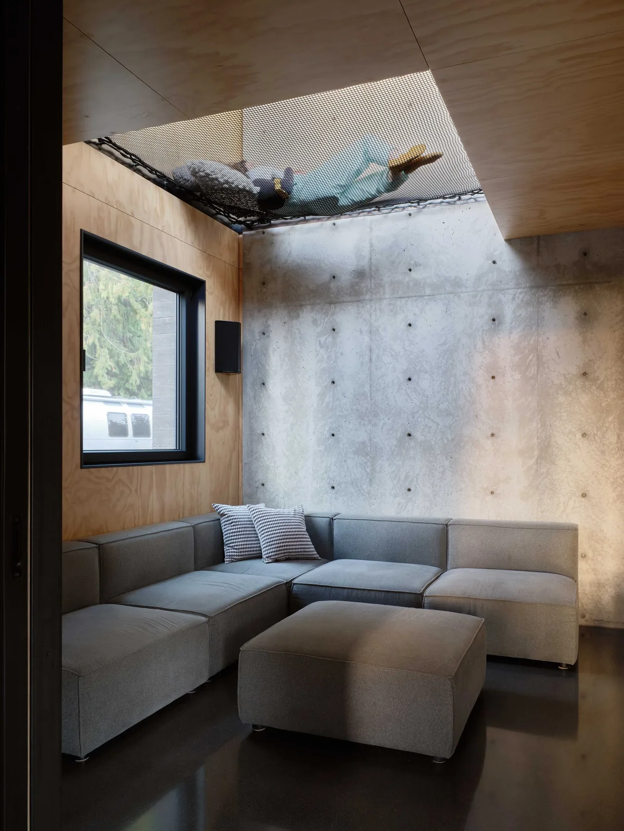 Living room features a gray sectional sofa, exposed concrete wall, plywood ceiling, and overhead mesh platform occupied by a person.
