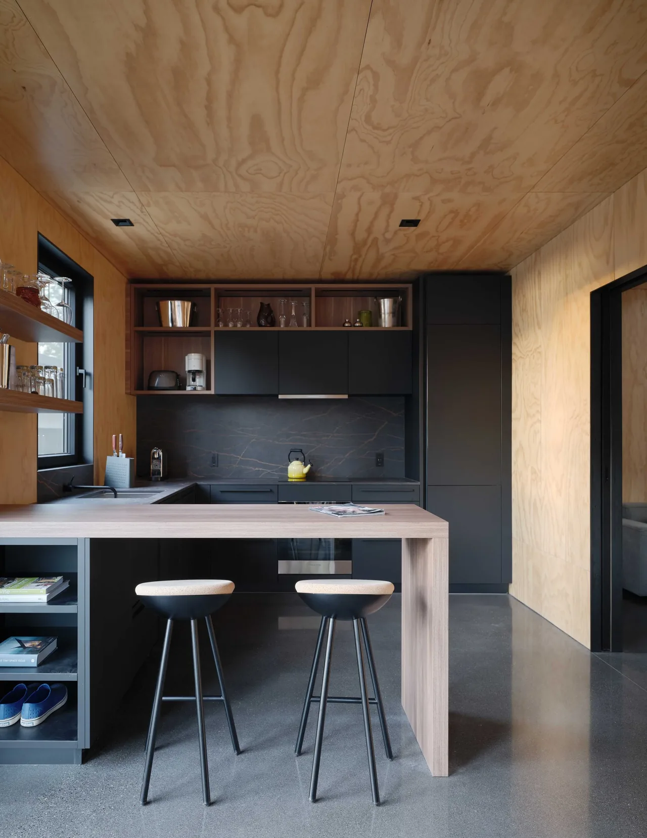 A modern kitchen with matte black cabinetry, light wood ceiling and countertop peninsula, and polished concrete floor.