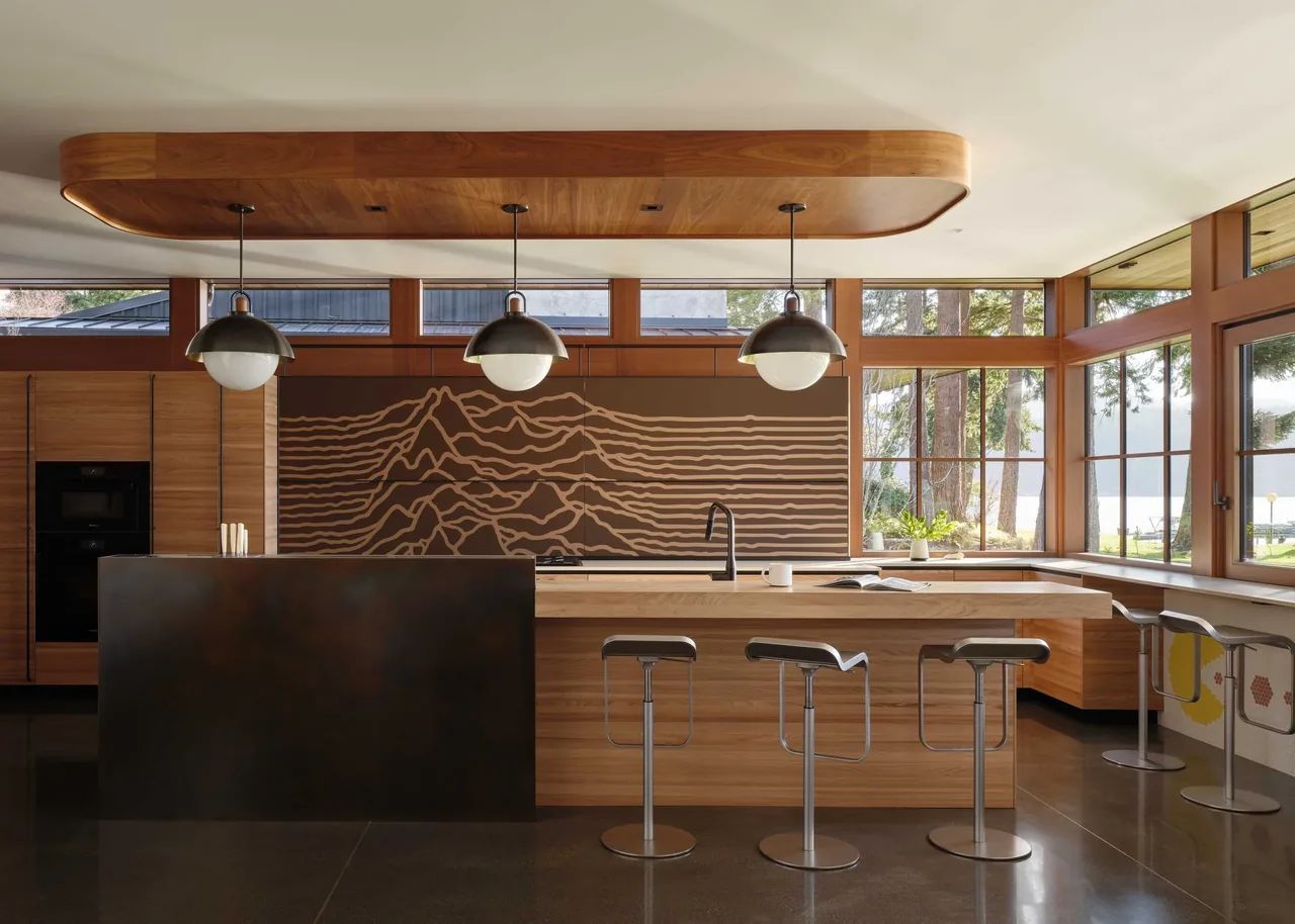 The Gamer HQ kitchen features wood grain cabinets, a large island, and corner windows with a lake view, designed by skB Architects.