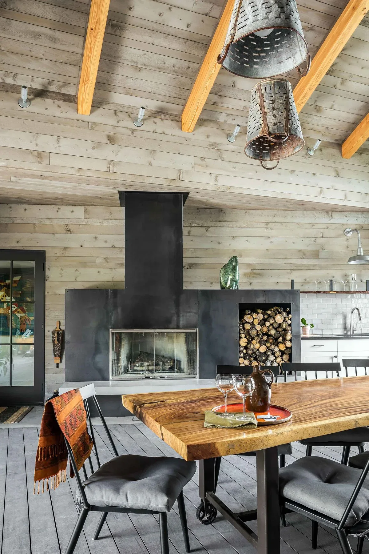 Dining area features exposed wood beams, metal basket pendant lights, and a fireplace with integrated wood storage at Roses Lake Residence.