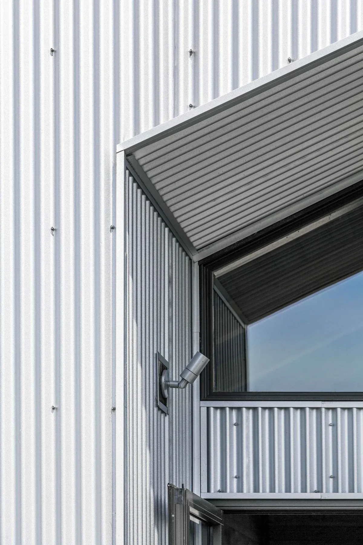 Corrugated metal siding and a contemporary black-framed window with an exterior spotlight at Roses Lake Residence.