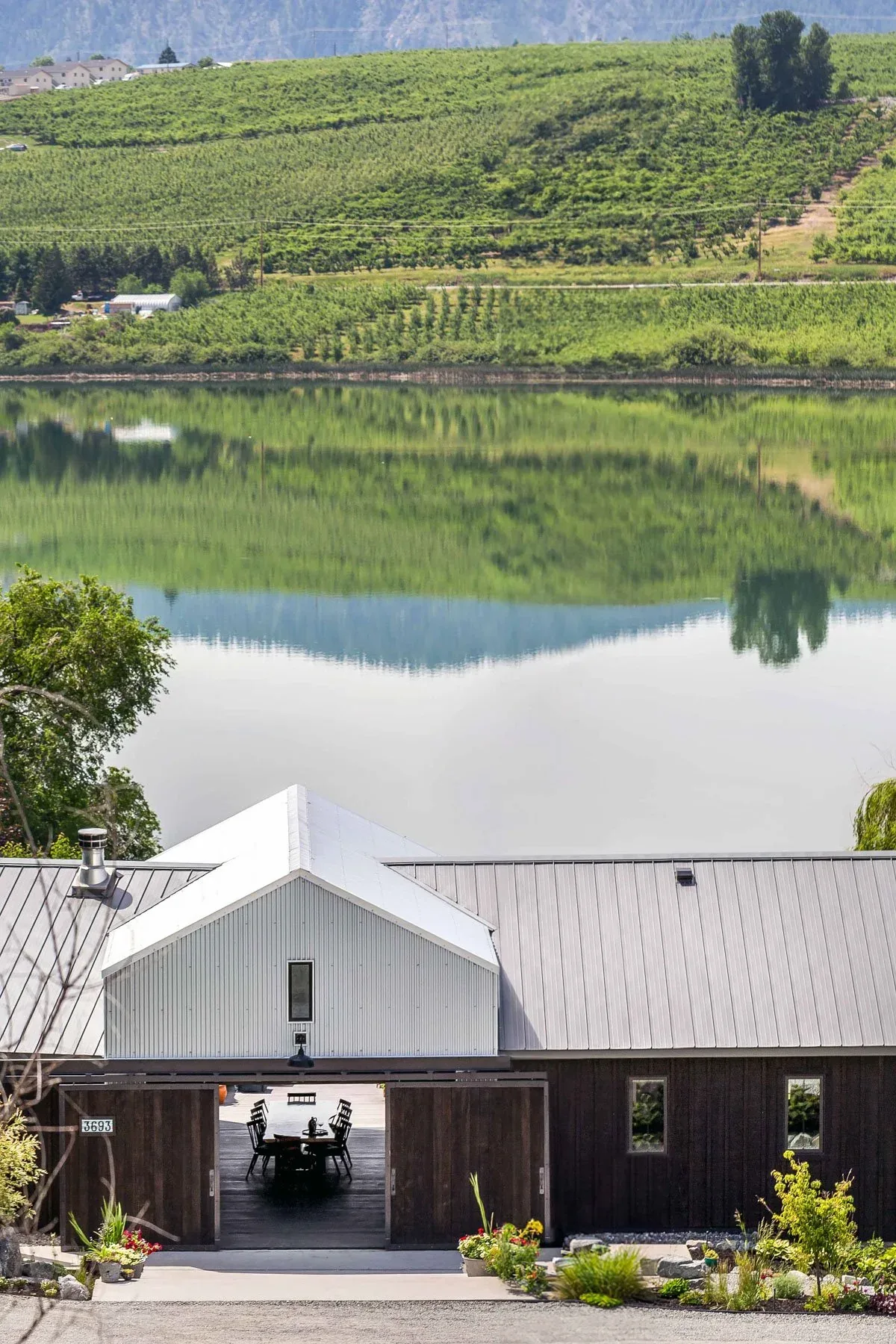 Barn-style building with metal roof and dark wood siding by a lake reflecting surrounding hills.