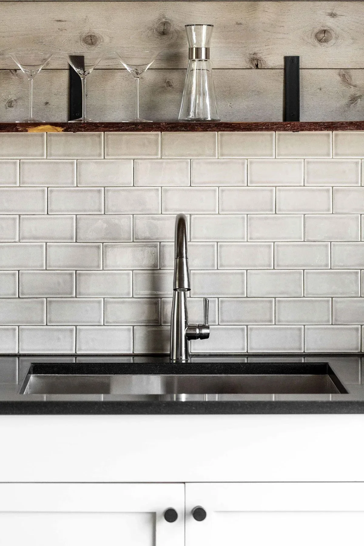 Kitchen sink area with stainless steel faucet and white subway tile backsplash beneath a rustic wood shelf with glassware.