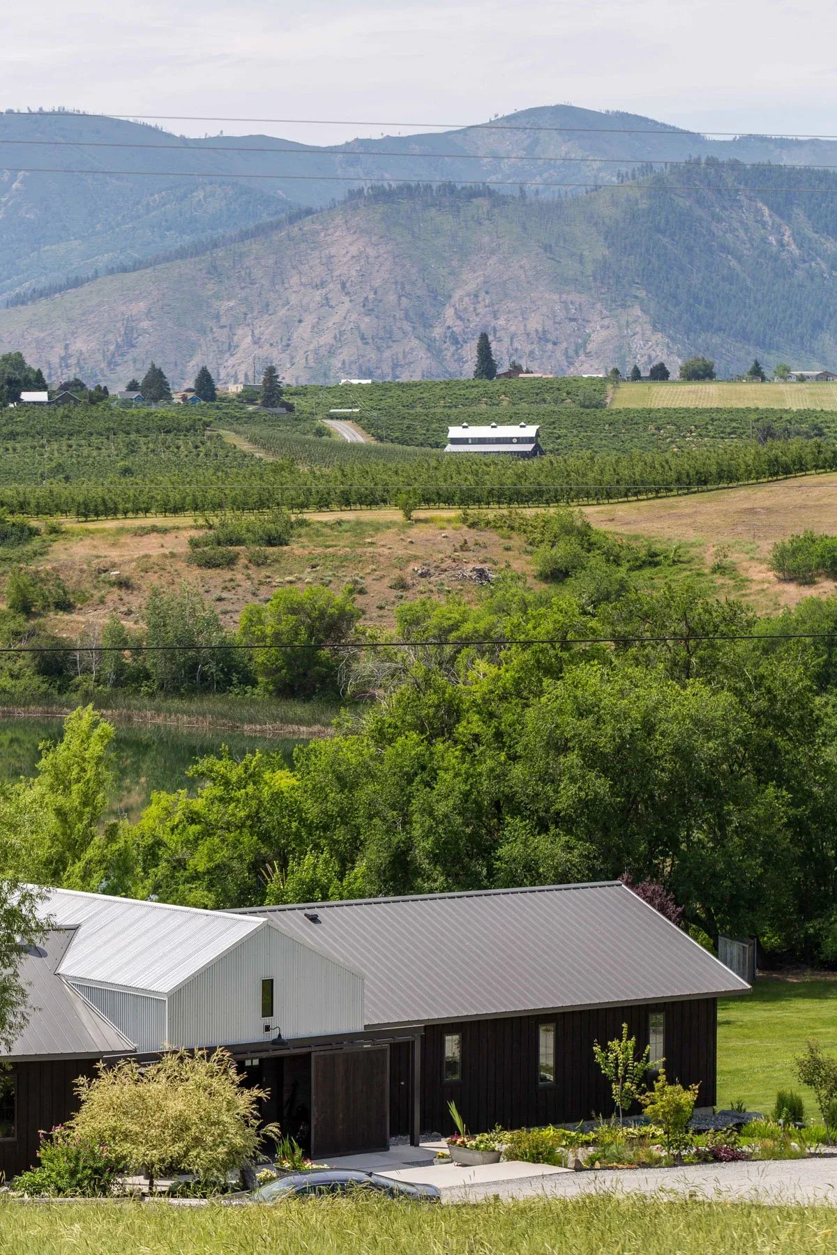 Barn-style residence with black siding and metal roof amidst farmland and distant mountains.
