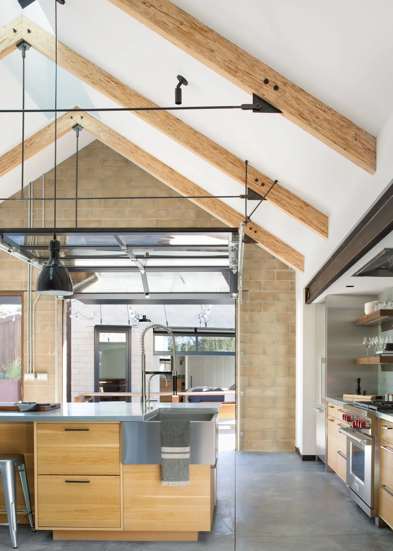 Modern kitchen features wood island, stainless steel appliances, exposed angled beams, and a glass garage-style door opening to the patio.