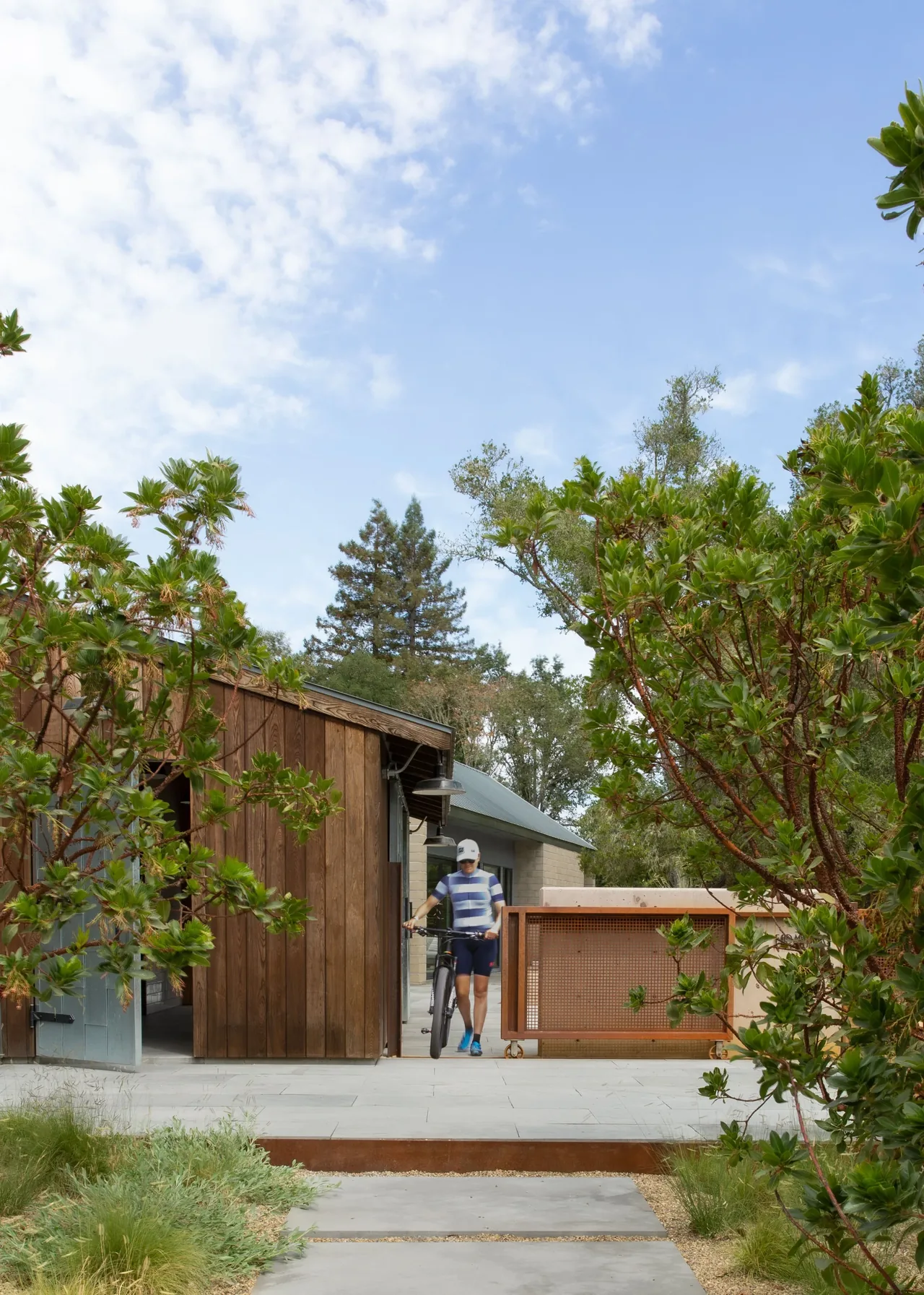 A man walks a bicycle alongside a wood-sided building with a concrete paved outdoor area and greenery.