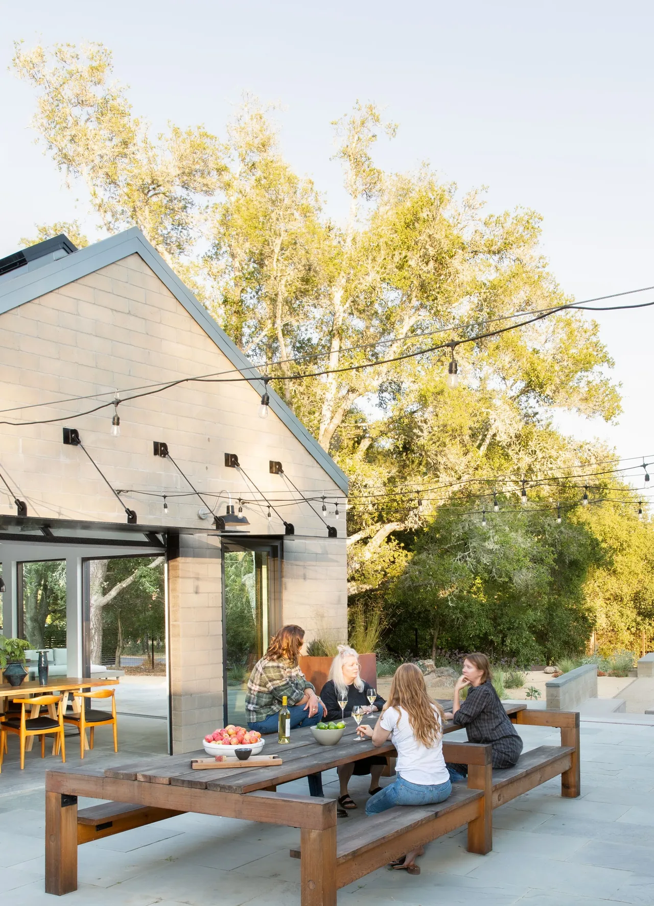 Outdoor dining area with a large wooden table and benches on a stone patio, next to a house with a gable roof and string lights overhead.