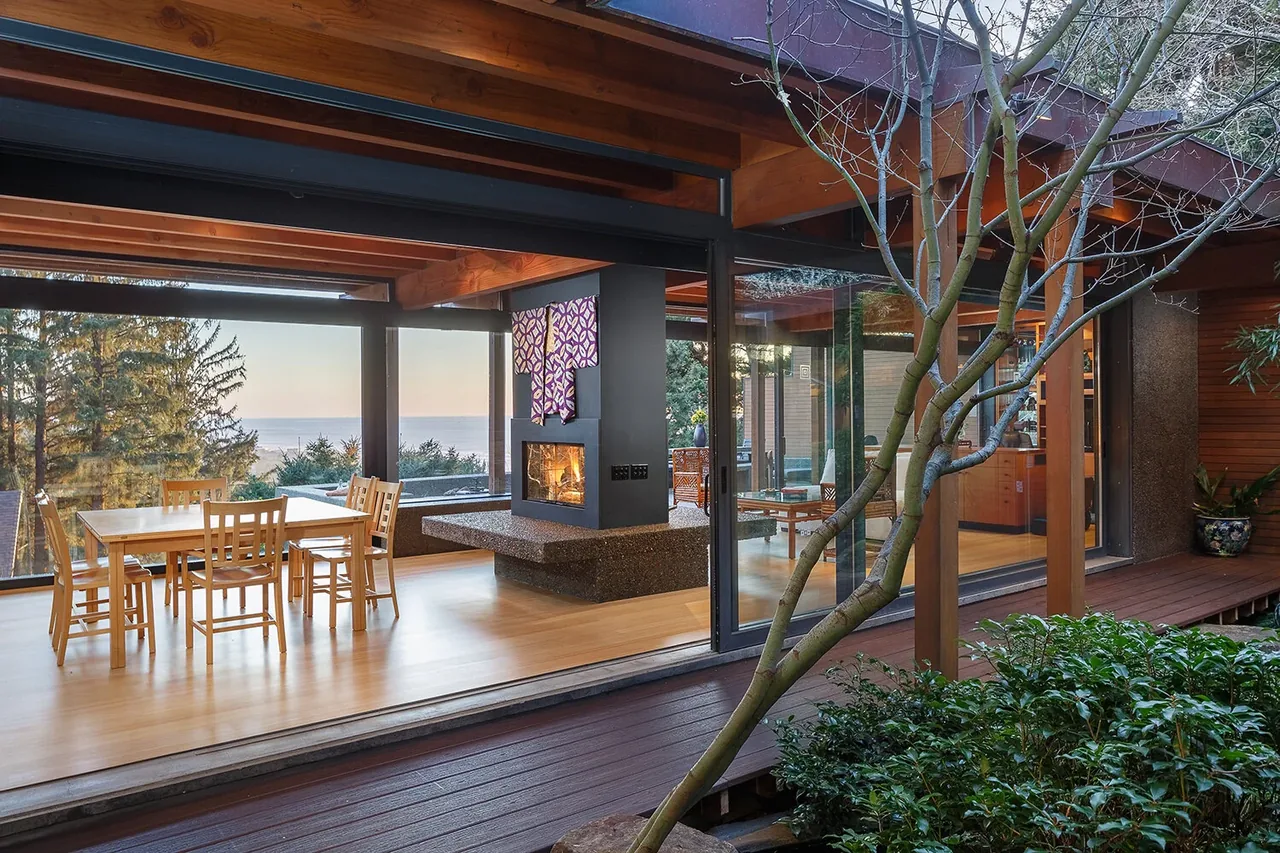 Dining area with wooden table and chairs features a central double-sided fireplace and large glass sliding doors opening to an outdoor garden deck.
