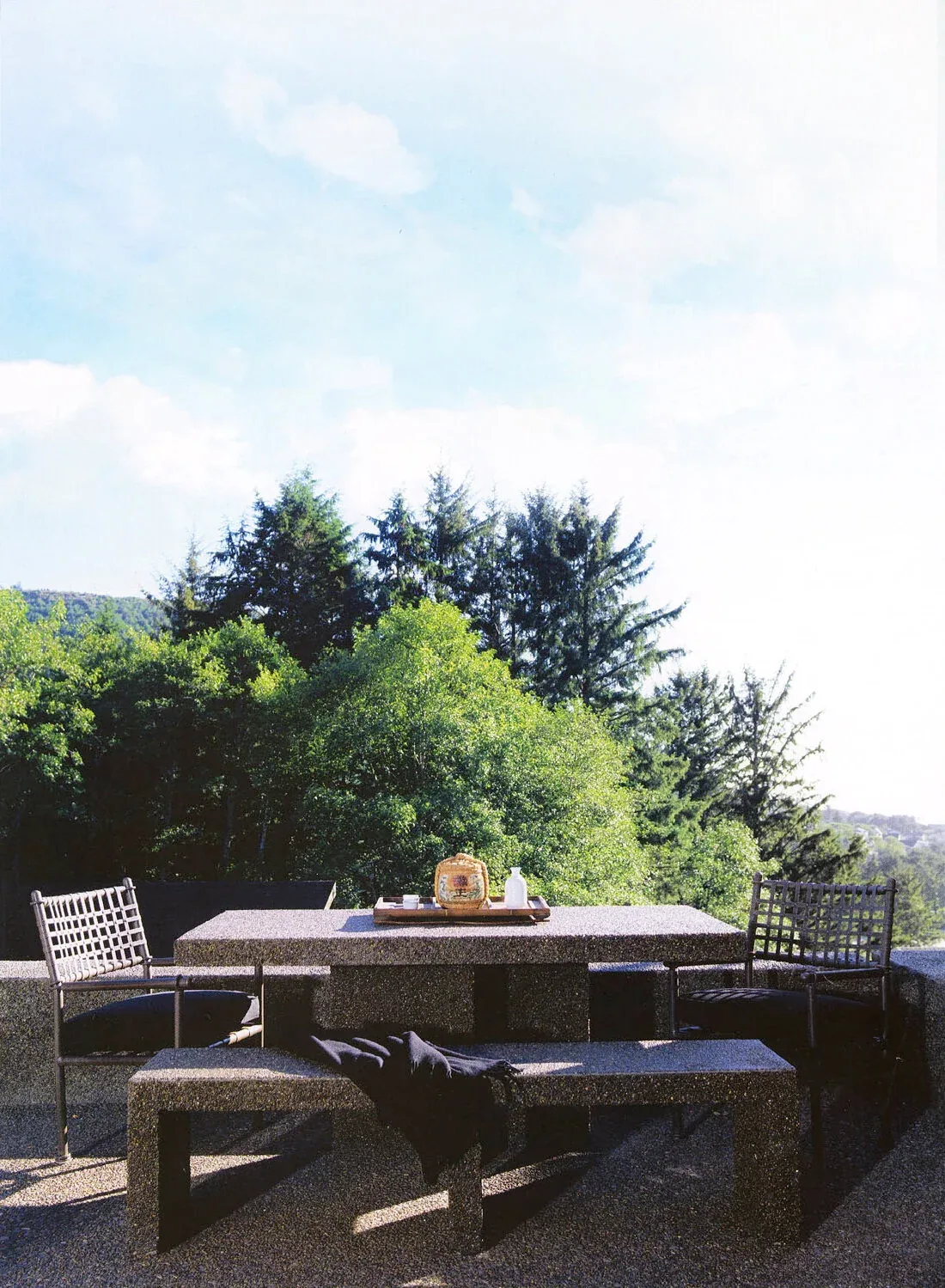 Outdoor dining area with a stone table, benches, and chairs overlooking a natural forested landscape and distant mountains.