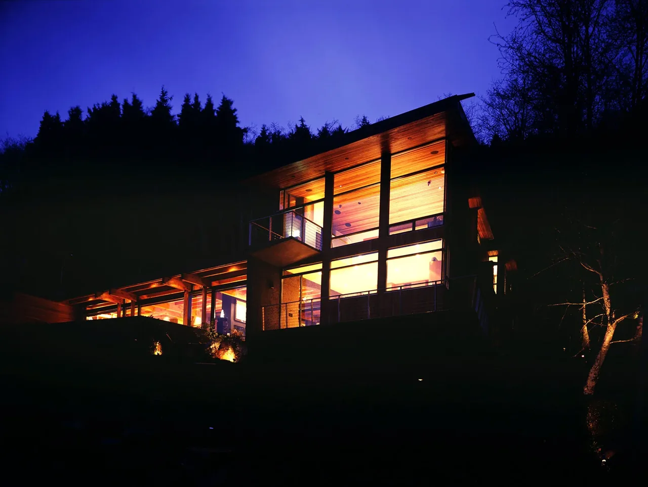 Night view of Horizon Hill House showing illuminated large windows and a sloped roof against a forest backdrop.