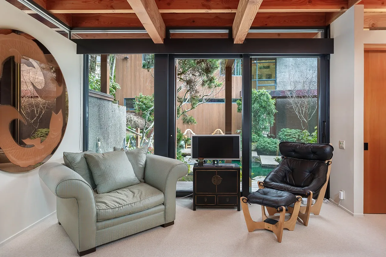 Living space with exposed wood ceiling beams and large black-framed windows facing a green courtyard with water features.