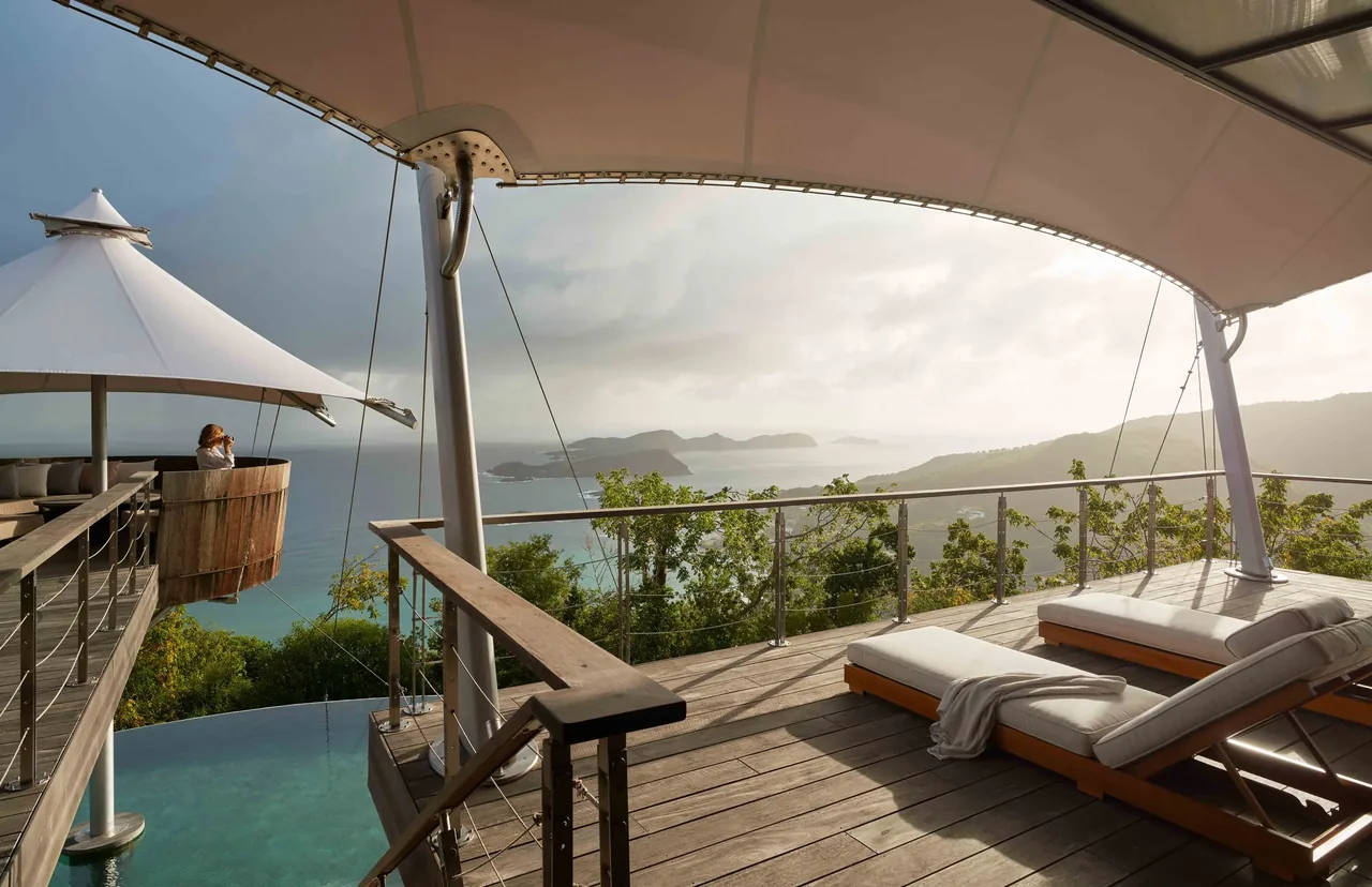 The SAIL HOUSE features a wooden deck with sun loungers and triangular shade sails overlooking a coastal landscape from an infinity edge pool.
