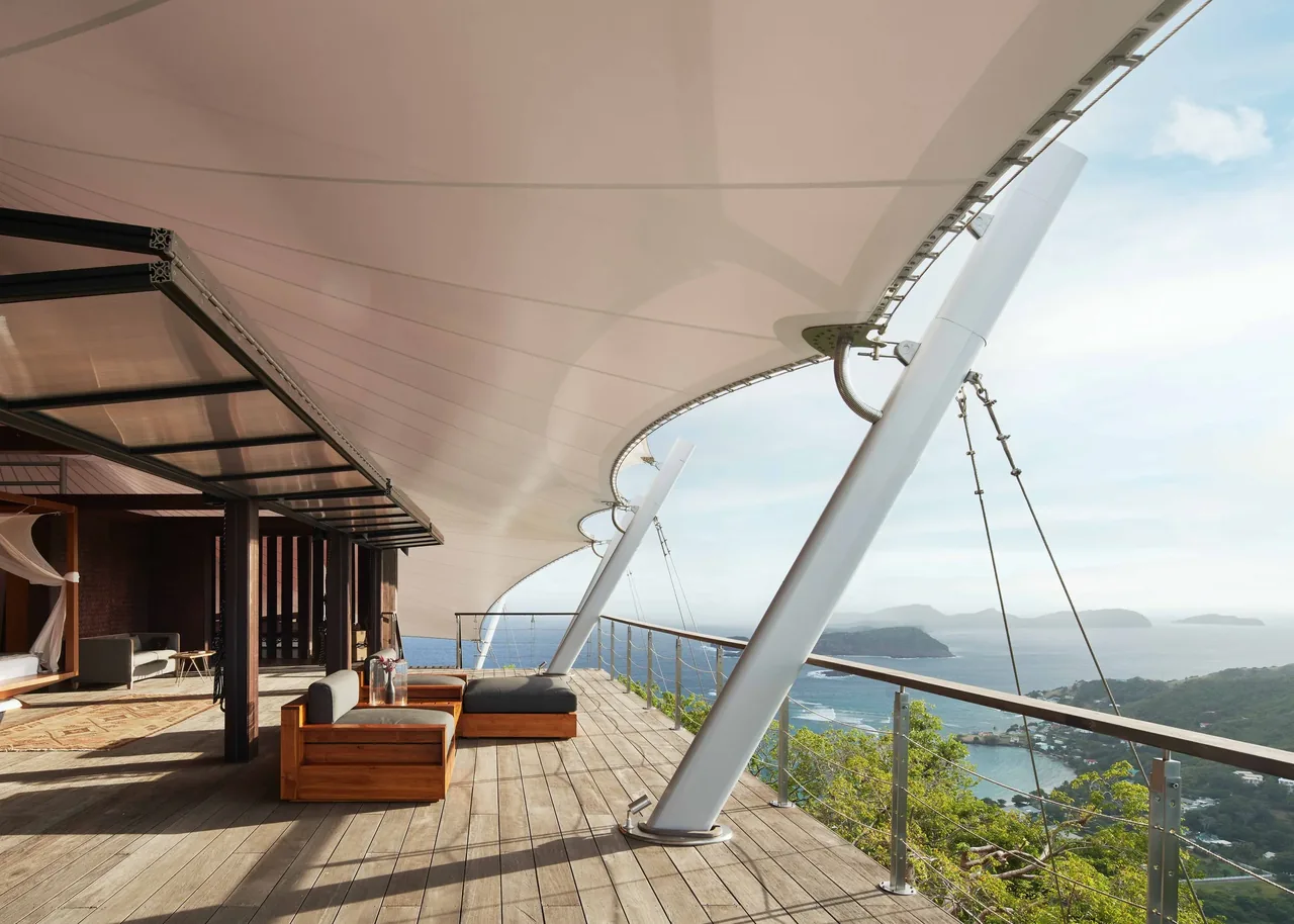 The outdoor living area features a wooden deck shaded by a large curved sail, offering coastal views and seating.