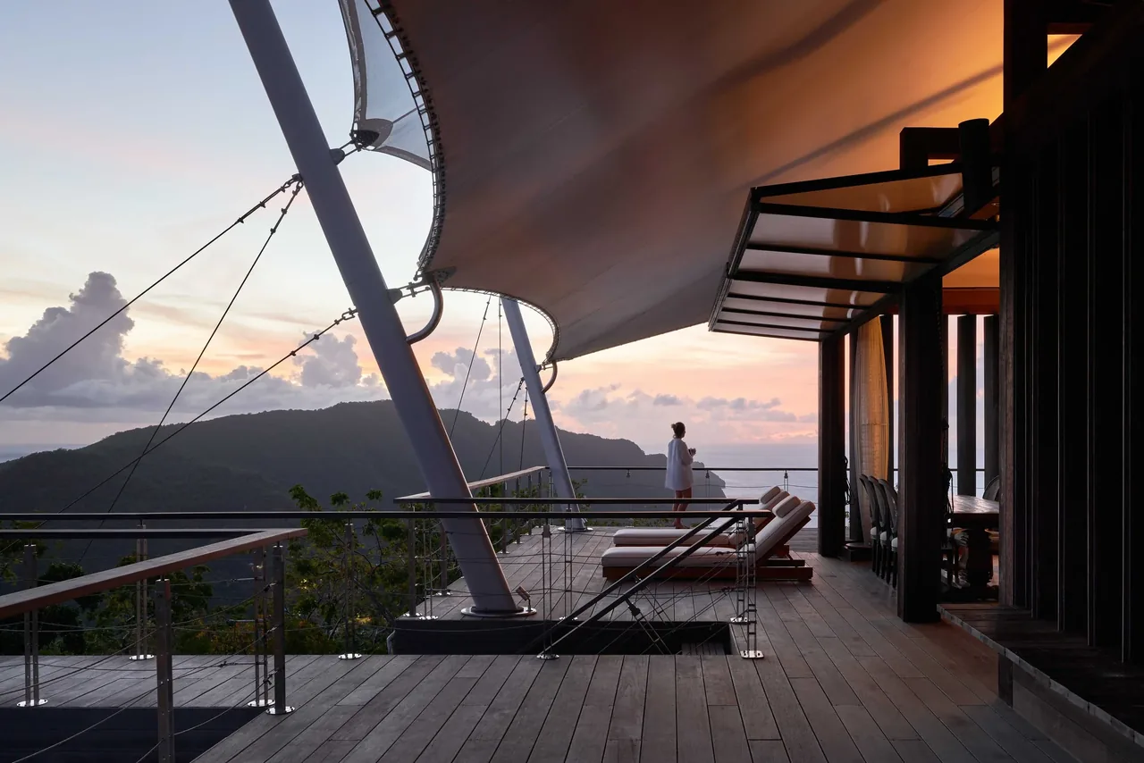 Outdoor wooden deck with triangular shade sails and seating overlooking a coastal view at sunset.