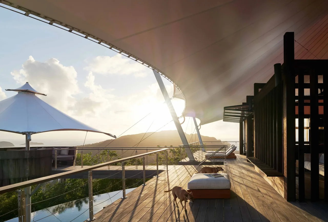 Wooden deck featuring triangular shade sails and lounge seating with panoramic outdoor views at sunset.