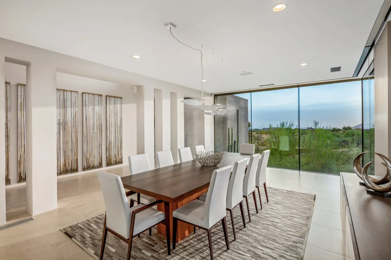 Dining area features a wood table with white leather chairs and large glass windows overlooking desert landscape.