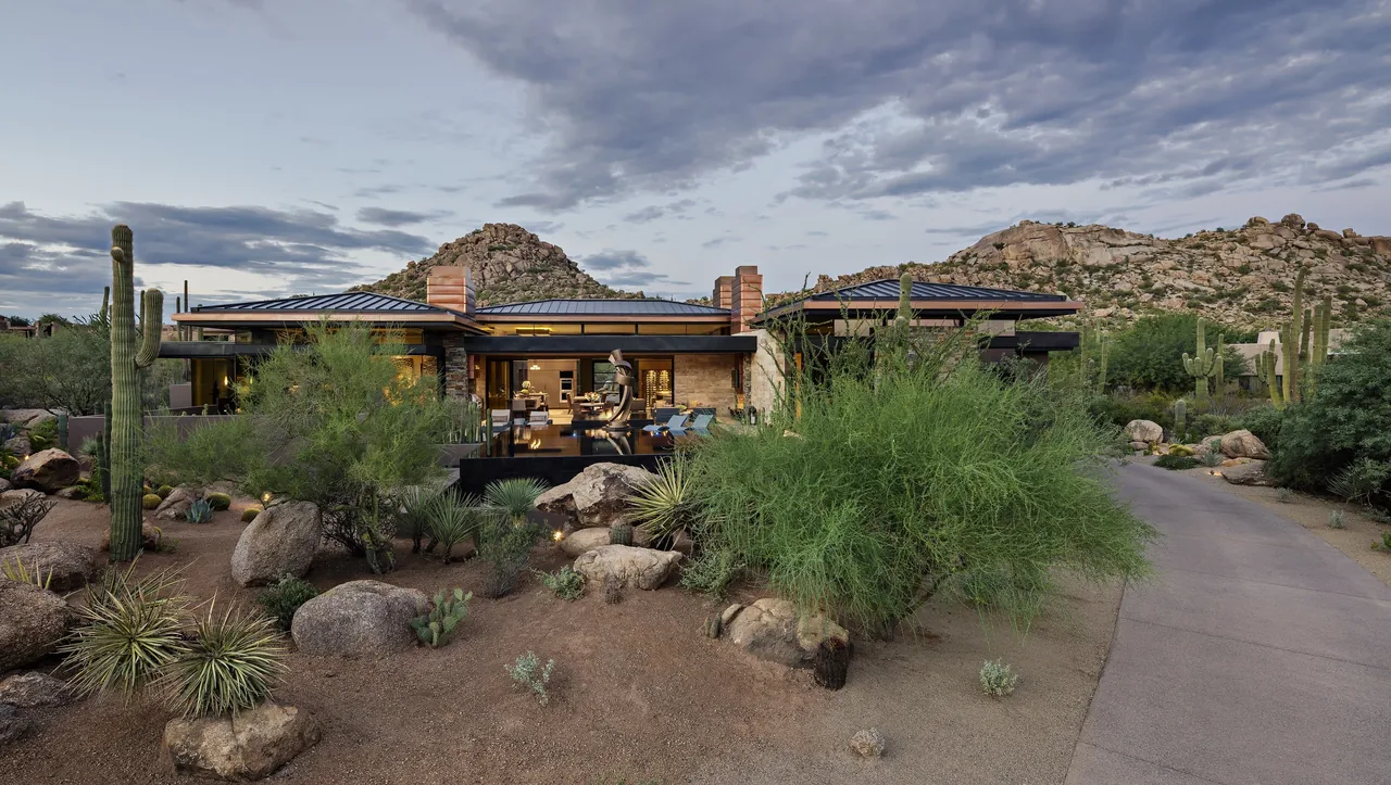 Silversmith Residence features desert landscaping and textured stone walls with a flat roof, set against rocky hills at sunset.