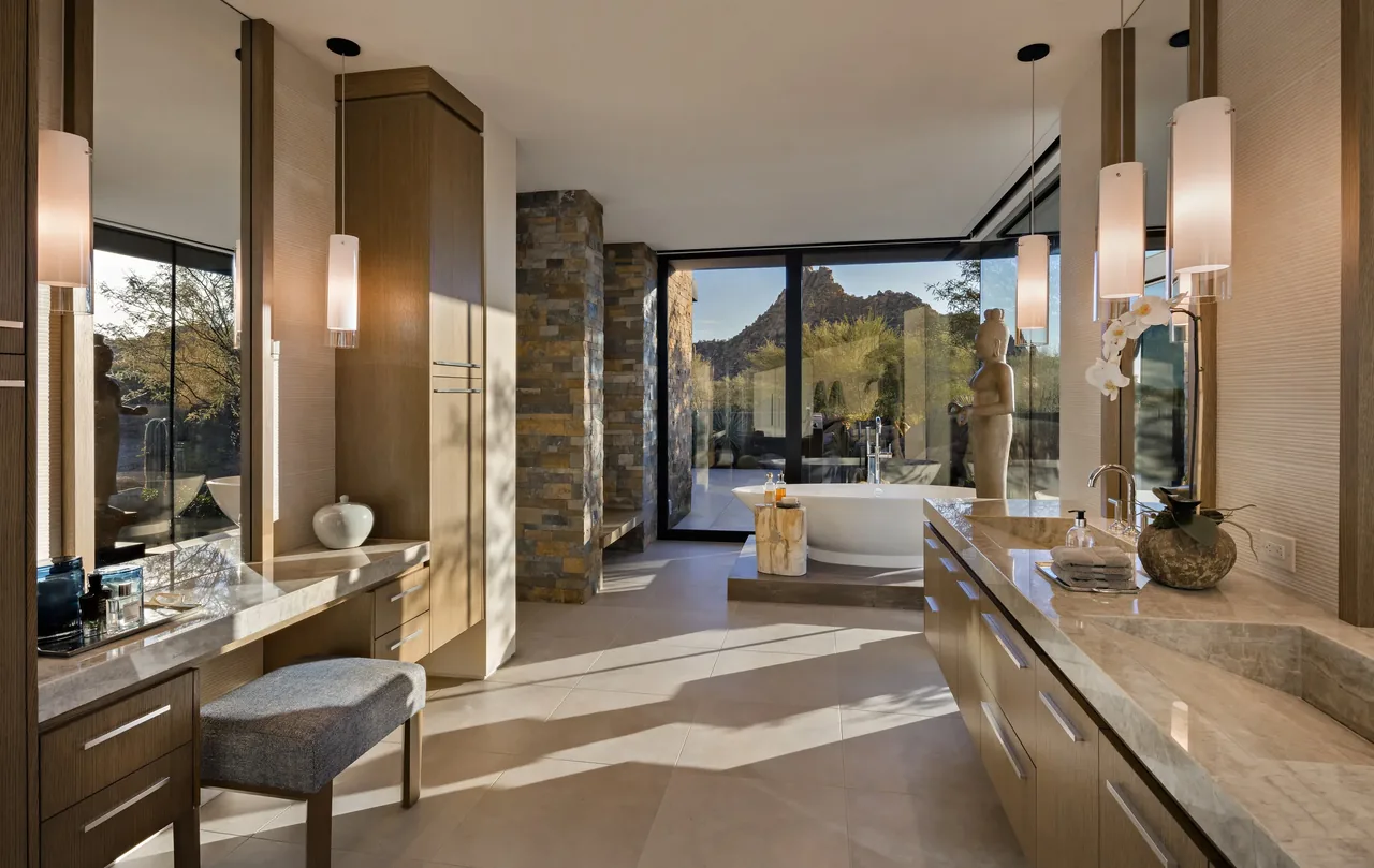 The Silversmith Residence bathroom features dual vanities with stone tops, pendant lighting, and a freestanding tub by large windows overlooking desert landscaping.