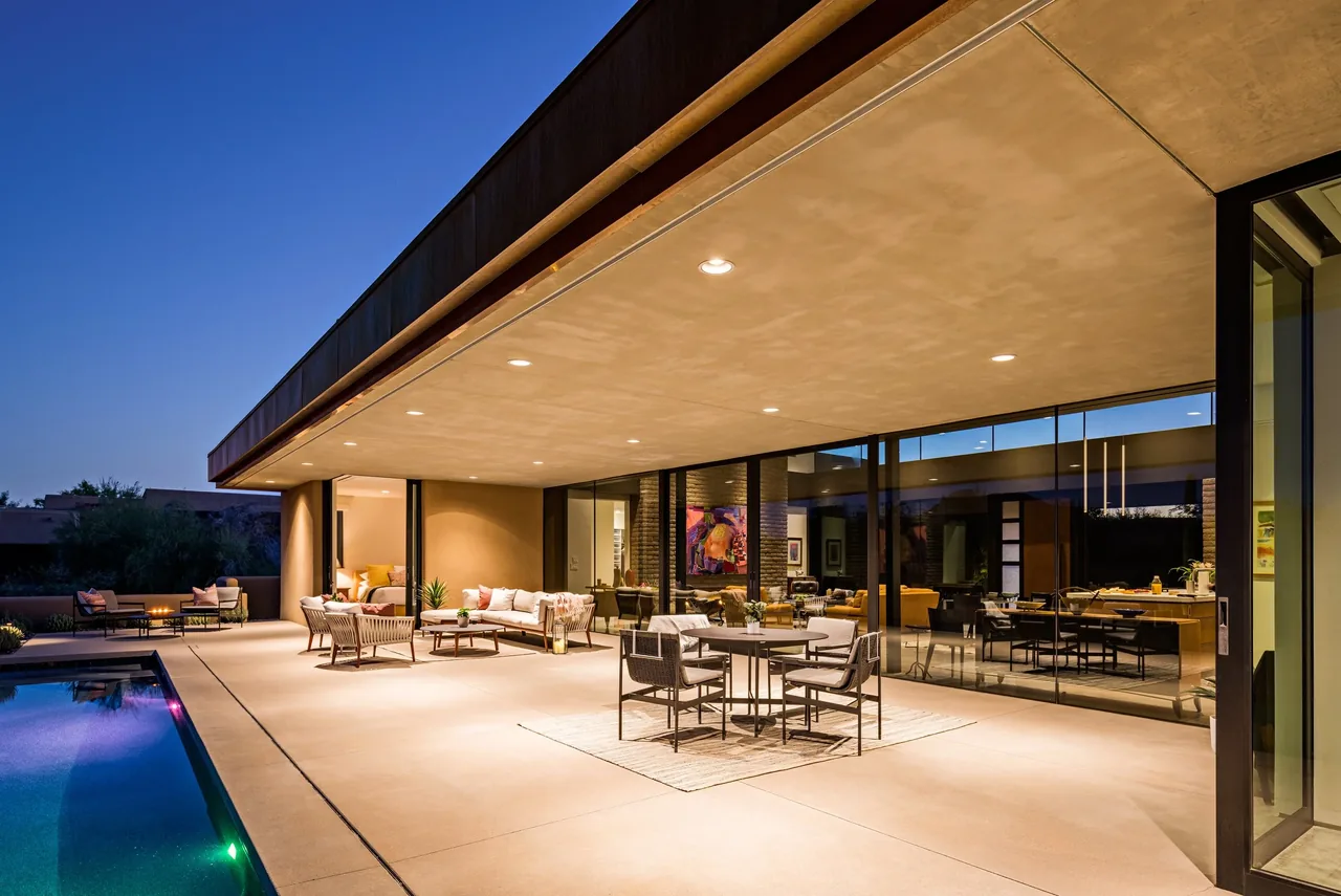 The Shea-Naito Residence features a covered patio with seating and dining areas adjacent to a rectangular pool at sunset.
