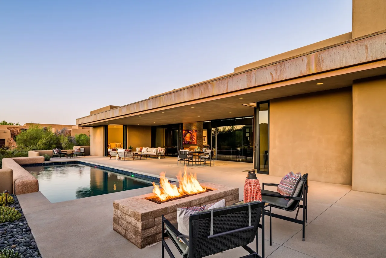 The Shea-Naito Residence features a rectangular fire pit with desert landscaping and a covered patio adjacent to a rectangular pool.