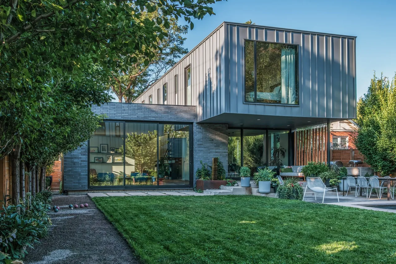 THE LOCUST HOME features dark brick, vertical metal siding, large window panels, and a wooden porch surrounded by landscaping.