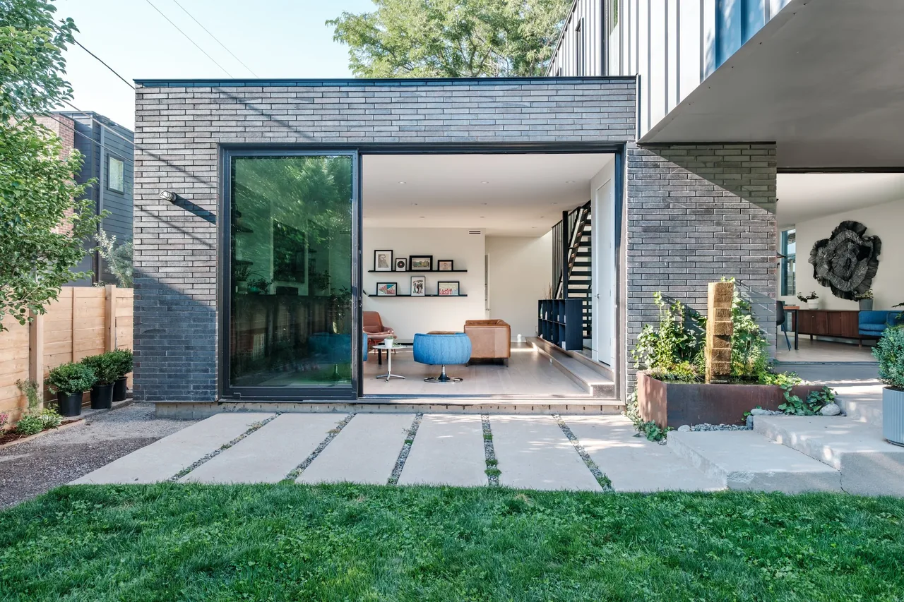 The rear exterior of THE LOCUST HOME features dark brick facade, sliding glass door, and concrete walkway leading to a modern living space.